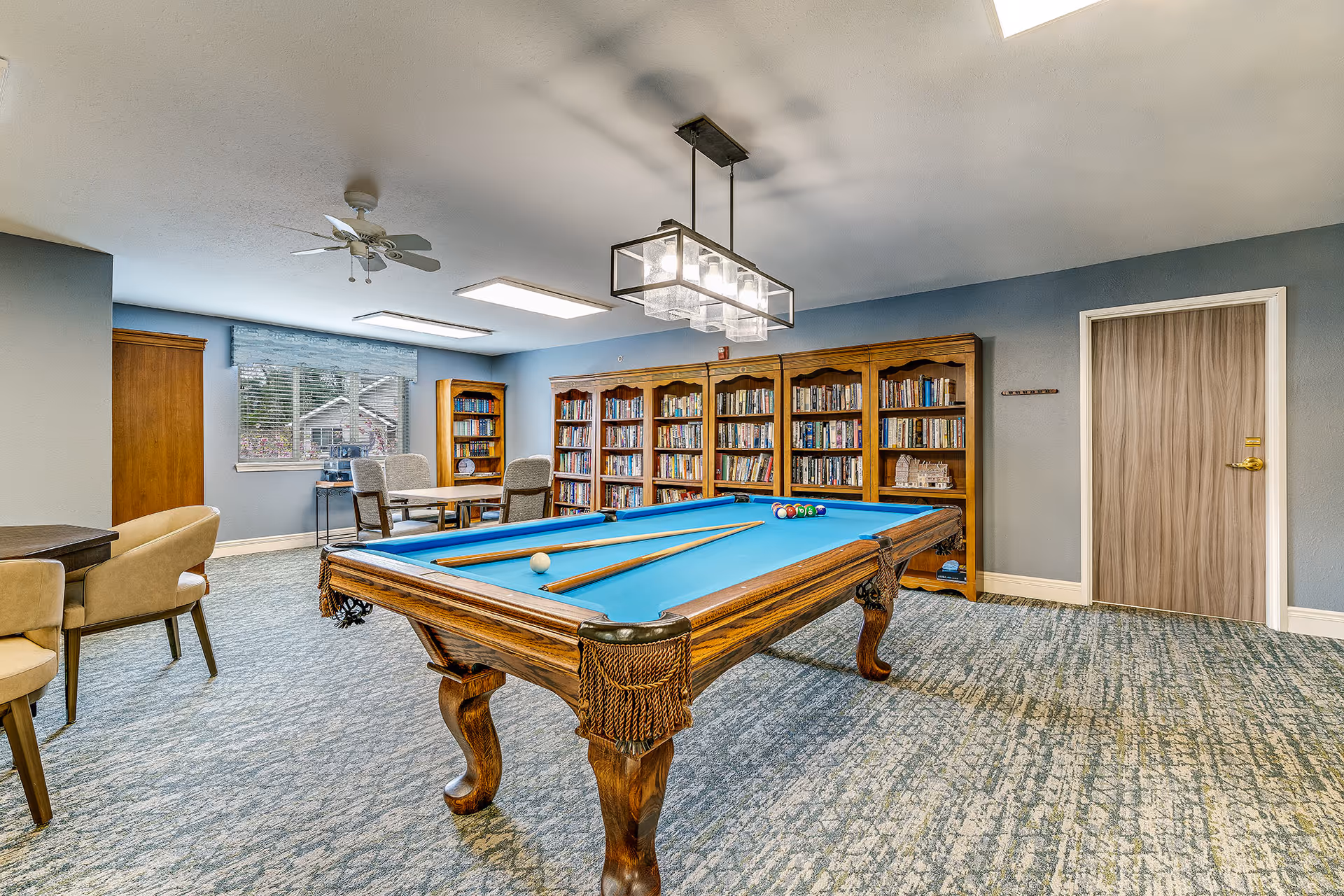 A recreational room with a blue felt pool table in the center, two pool cues and balls arranged on it. Behind the pool table, there are wooden bookshelves filled with books. To the left, there are tables with chairs and a window with blinds letting in natural light. The walls are painted light blue, and the carpet has a patterned design. A ceiling fan and a modern rectangular light fixture hang from the ceiling.