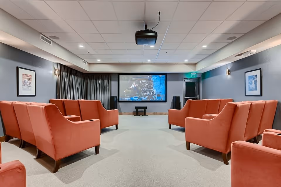 A small theater room with two rows of orange upholstered armchairs facing a large screen on the wall. The room has gray walls with framed pictures, a ceiling-mounted projector, and recessed lighting. There is an exit door to the right of the screen.