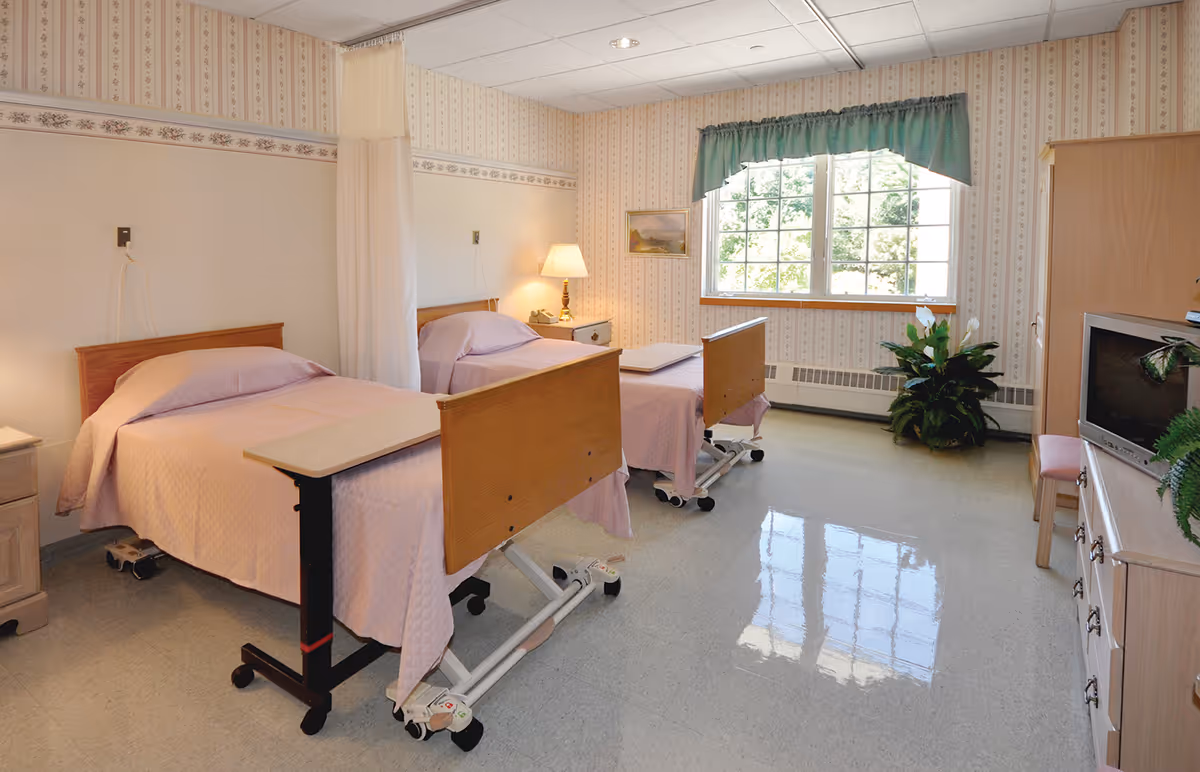 A bright shared patient bedroom with two adjustable beds, bedside tables, a large window with a valance, and a TV cabinet.