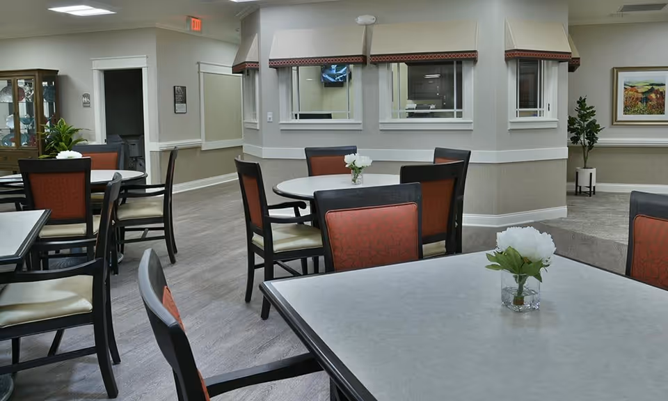 Dining area with multiple round and square tables, upholstered chairs, small floral centerpieces, and a serving window/desk area.