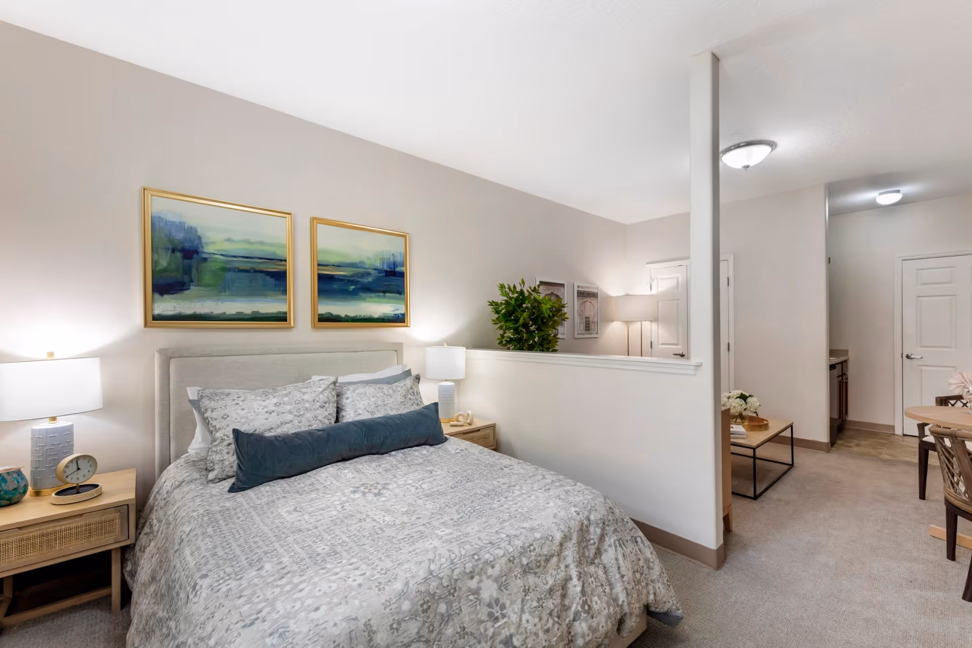 Furnished bedroom with a bed, bedside tables and lamps, artwork above the headboard, and a half wall opening to a living/dining area.