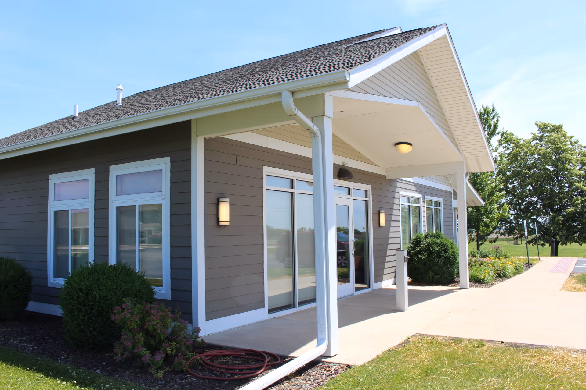 Exterior view of a single-story building with gray siding and white trim, featuring large windows and a covered entrance with a concrete walkway. There are bushes and greenery around the building under a clear blue sky.