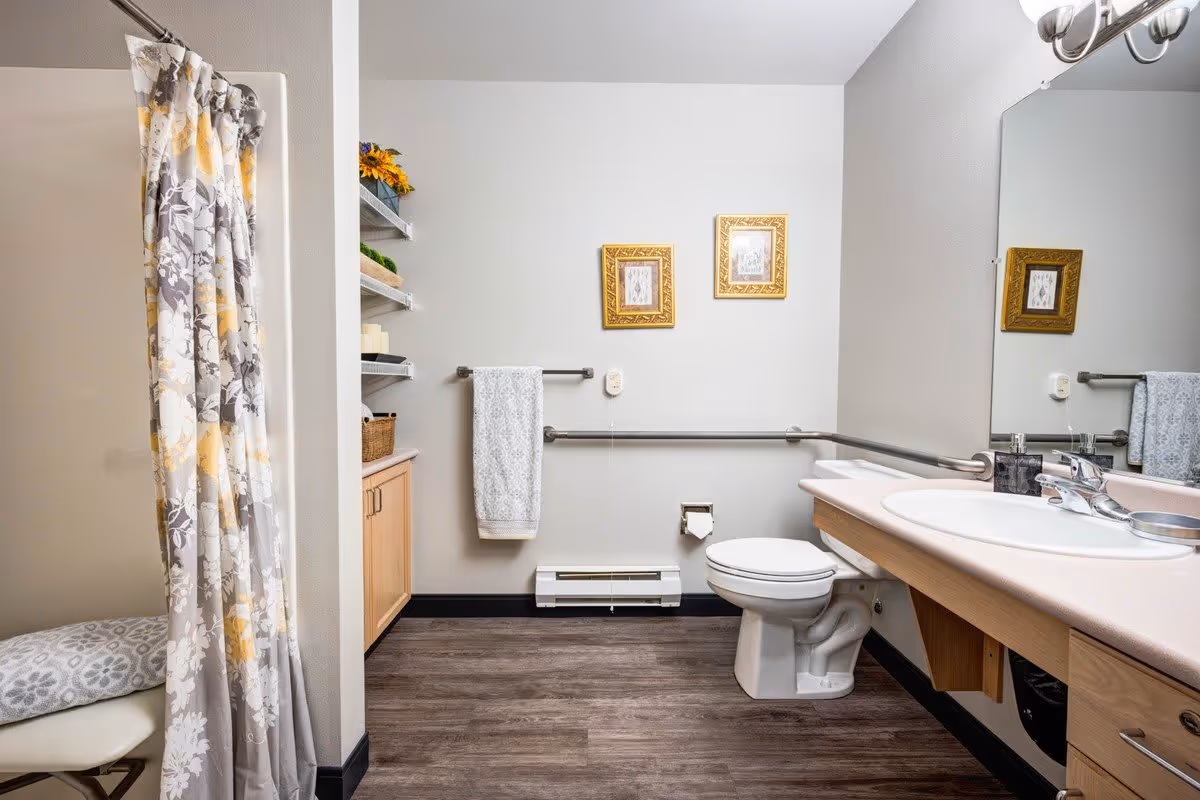 A clean and accessible bathroom with a toilet, sink with countertop, and a shower area with a floral-patterned curtain. The bathroom has wooden flooring, grab bars near the toilet, a towel hanging on a bar, two framed pictures on the wall, and shelves with decorative items and towels.
