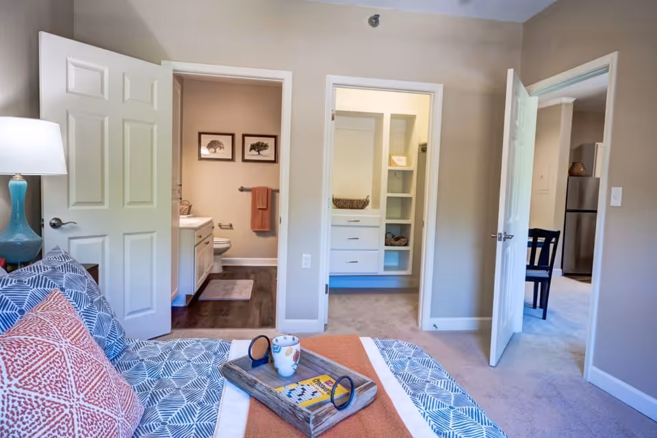 A bedroom with a bed featuring patterned bedding and orange pillows, a bedside table with a blue lamp, and a tray holding a mug and a book. The room has beige walls and carpeted floor. Two open doors reveal a bathroom with a toilet and sink, and a closet with shelves and drawers. Another open door leads to a dining area with a table and chairs and a stainless steel refrigerator.