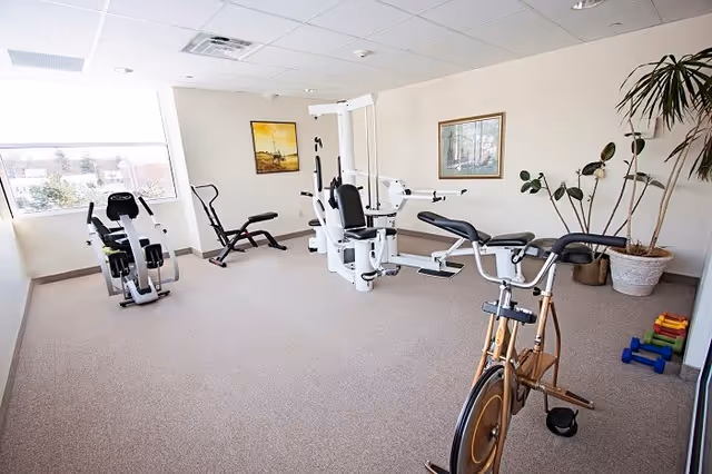 A bright exercise room with various fitness equipment including a stationary bike, rowing machine, multi-function weight machine, and dumbbells. The room has large windows letting in natural light, framed artwork on the walls, and a potted plant in the corner.