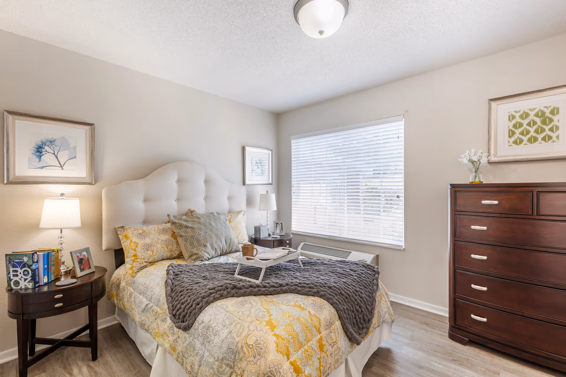 Sunlit bedroom with a tufted headboard, patterned bedding, bedside tables with lamps, and a tall wooden dresser.