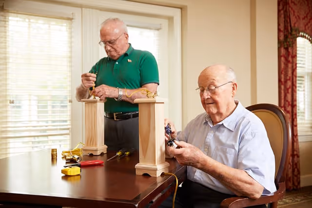 Two elderly men working on assembling or repairing wooden lamp bases with electrical cords and tools on a wooden table in a well-lit room with large windows and curtains.