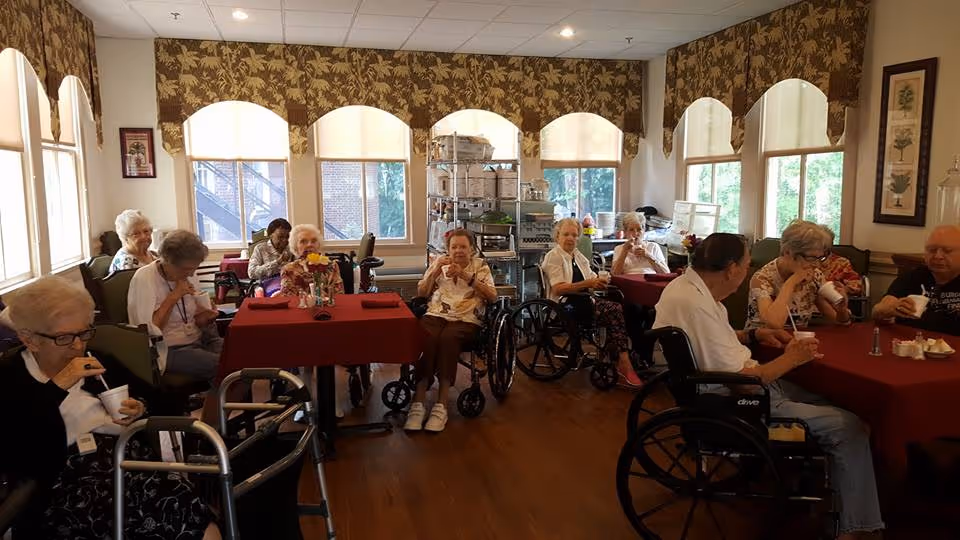 A group of elderly residents sitting around tables with red tablecloths in a well-lit room with large windows and floral curtains. Some residents are in wheelchairs and others use walkers. They appear to be enjoying drinks and socializing in a communal dining or activity area.