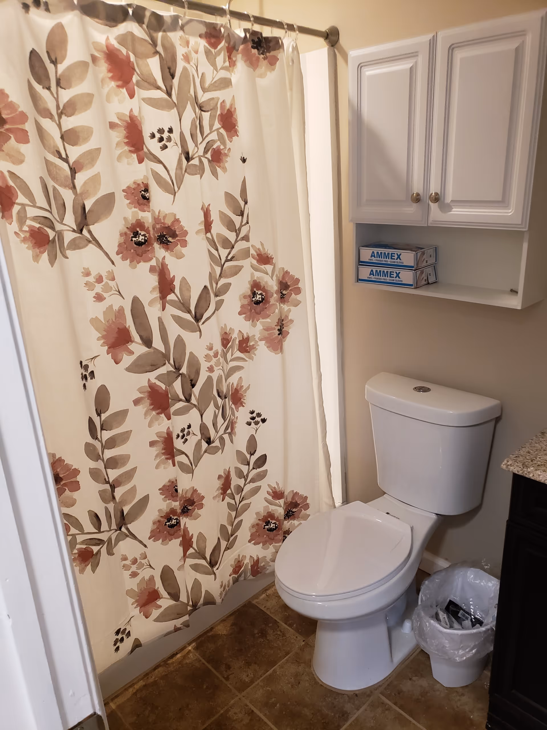 A bathroom with a white toilet, a floral-patterned shower curtain in shades of pink and brown, a white cabinet mounted on the wall containing two boxes of AMMEX gloves, a small trash can with a plastic liner, and a granite countertop partially visible on the right side.