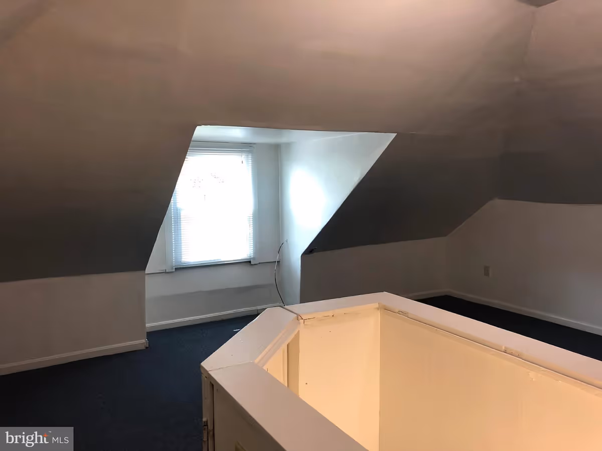 An empty attic room with slanted ceilings, a window with blinds letting in light, dark carpeted floor, and a white railing around a stairwell opening.