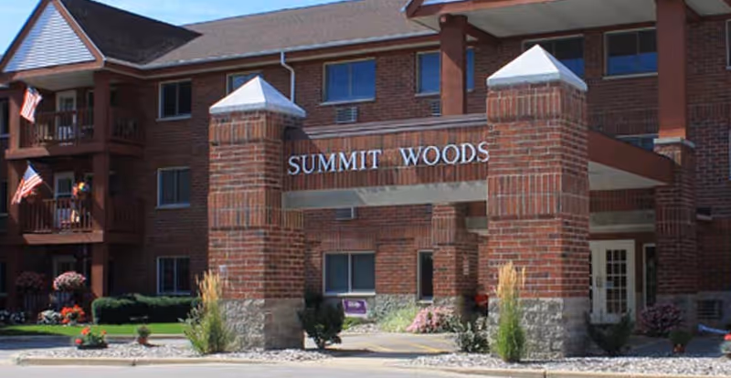 Front entrance of a brick multi-story building with a porte-cochere displaying the sign 'SUMMIT WOODS', landscaping, and balconies with American flags.