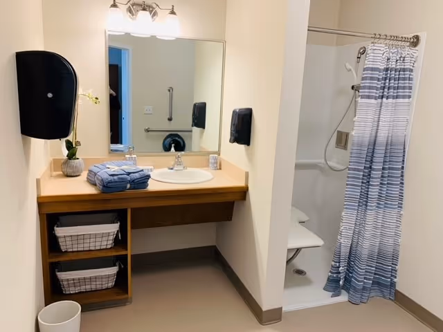 A clean bathroom with a wooden vanity holding a sink, a mirror above it, and a small potted plant and folded towels on the counter. To the right is a walk-in shower with a white fold-down seat and a blue and white striped shower curtain. There are two wire baskets on shelves under the vanity and a white trash bin on the floor.
