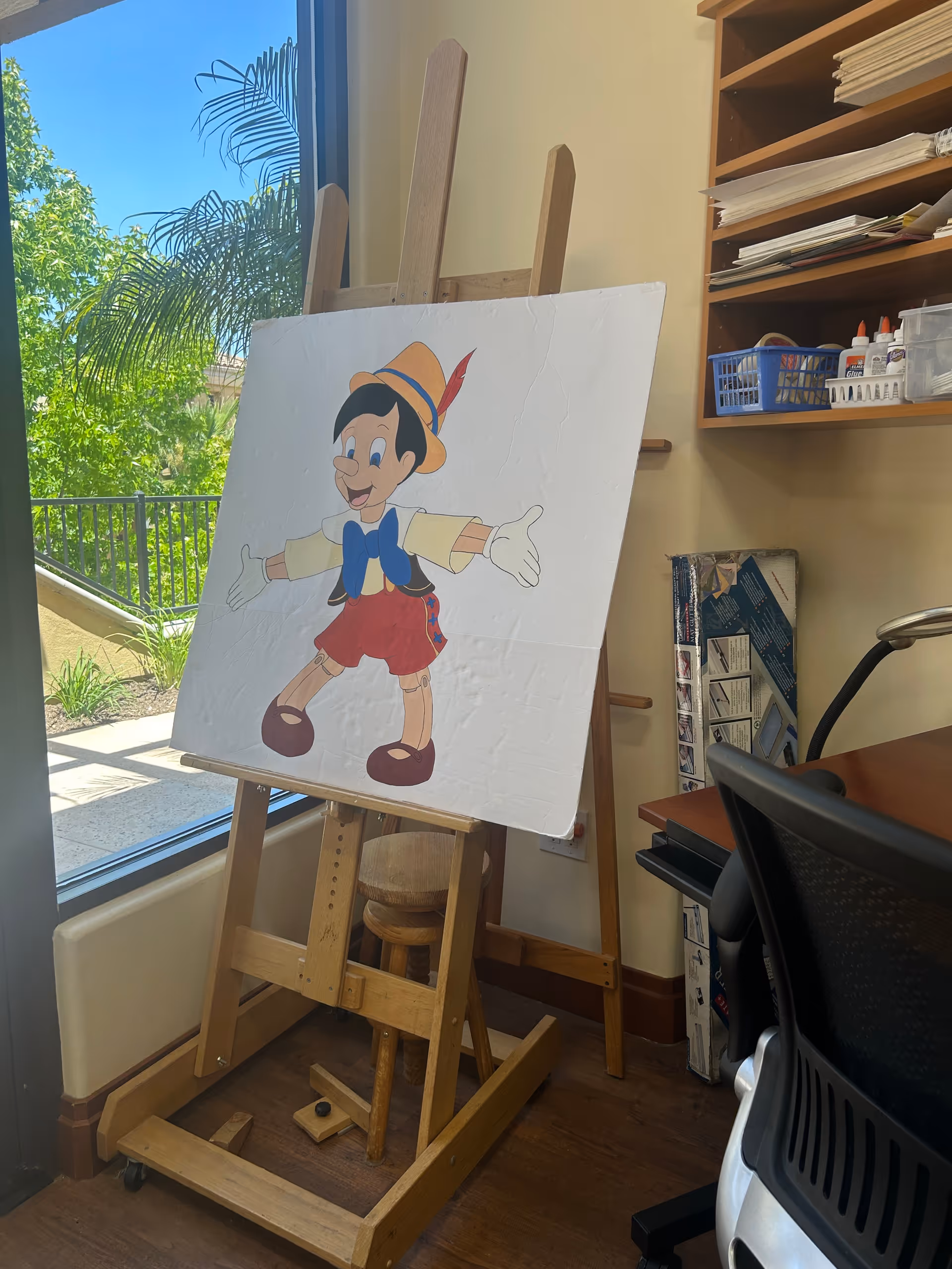 A painting of Pinocchio on a wooden easel in a room with a large window showing green trees outside. The room has a wooden floor, a black office chair, a wooden stool, and shelves with various supplies including glue bottles and papers.