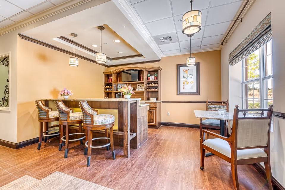 A cozy interior space featuring a wooden bar with three cushioned bar stools, pendant lights hanging from the ceiling, a wooden cabinet with shelves holding decorative items, and a small dining area with a table and two chairs near a window with a patterned valance.