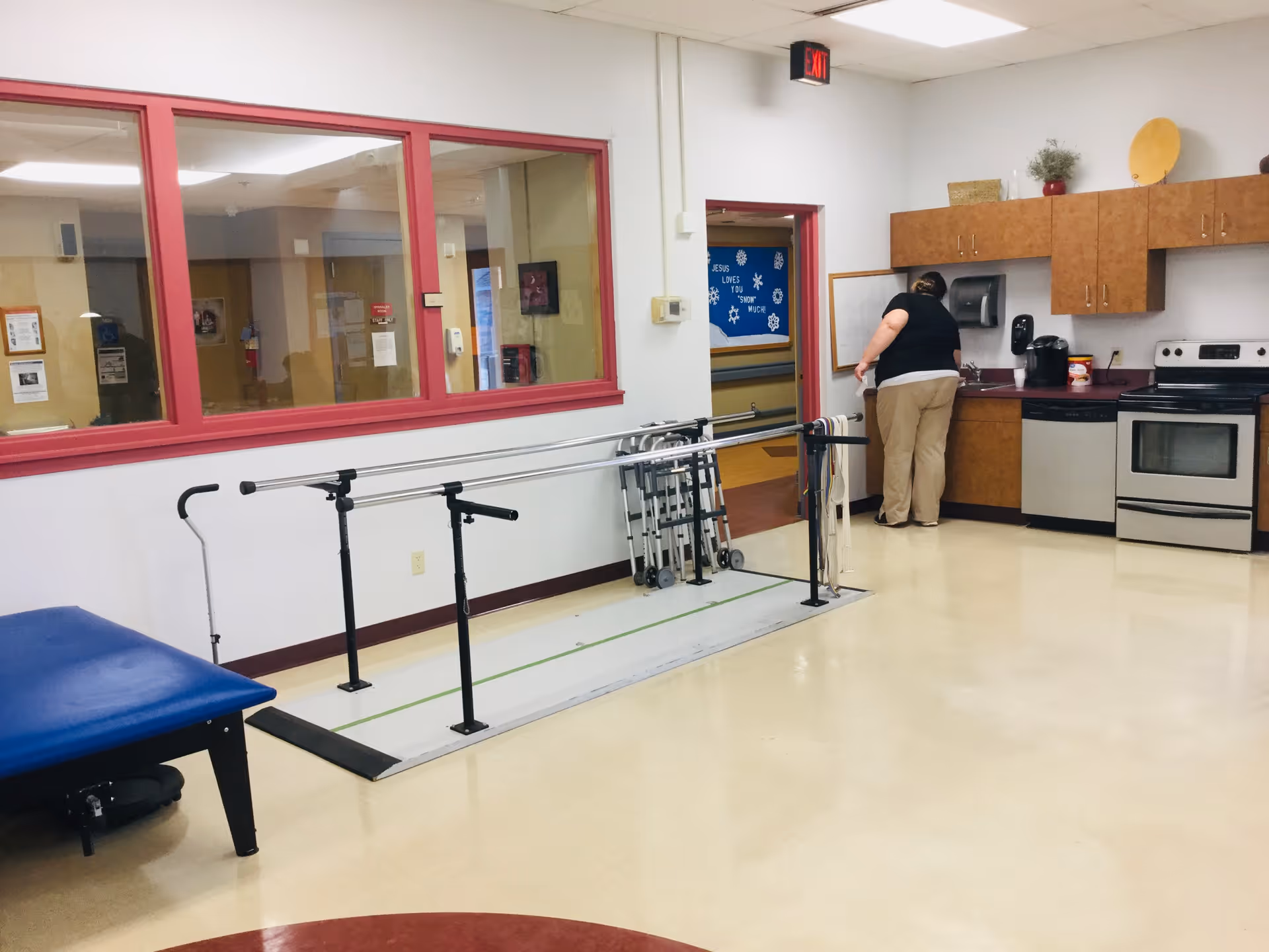 A room with a kitchen area on the right side including a stove, dishwasher, and cabinets. A person is standing near the counter. In the center of the room, there are parallel bars used for physical therapy or walking assistance. Several walkers are lined up against the wall. There is a blue padded bench on the left side and large windows with red frames on the left wall.