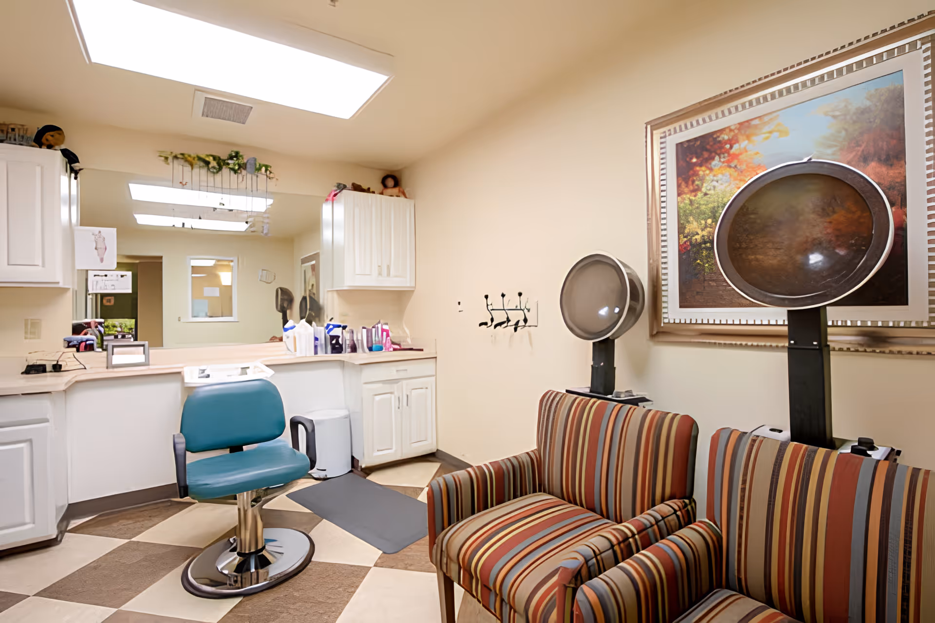 Interior of a salon area in a senior living facility featuring a teal salon chair in front of a large mirror, white cabinets, and two striped armchairs with hair dryers above them. A framed colorful landscape painting hangs on the wall.
