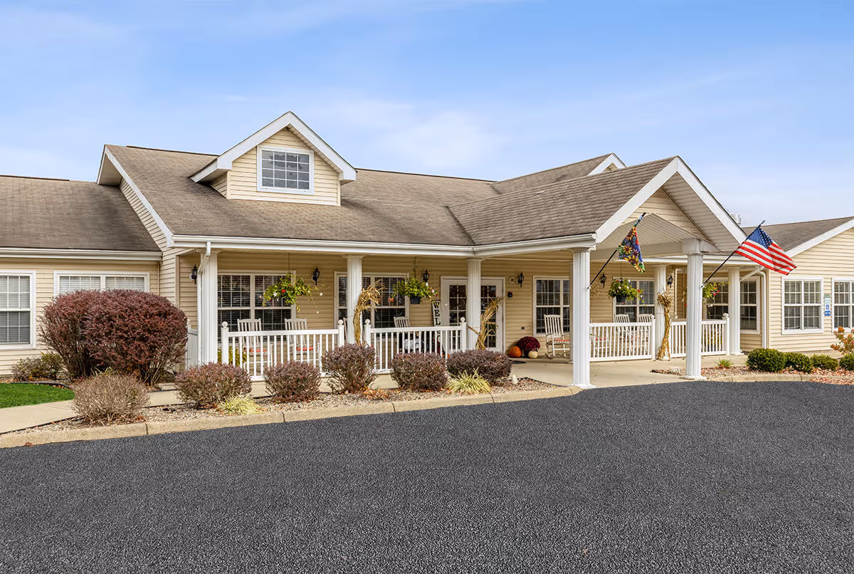 Exterior front view of a single-story building with beige siding and a covered porch supported by white columns. The porch has white railings, rocking chairs, hanging plants, and seasonal decorations including pumpkins and cornstalks. An American flag is displayed near the entrance. The building is surrounded by small bushes and a paved driveway.