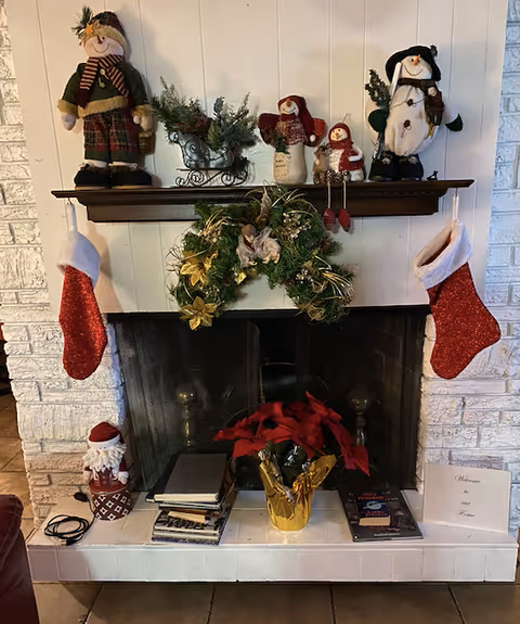 A cozy fireplace decorated for Christmas with two red stockings hanging on each side of the mantel. The mantel features festive decorations including a snowman figurine, a small sleigh with greenery, and three small snowman dolls. A green wreath with gold accents hangs above the fireplace opening. Below, on the hearth, there is a poinsettia plant wrapped in gold foil, a small Santa figurine, several stacked books, and a card that reads 'Welcome to our Home'.