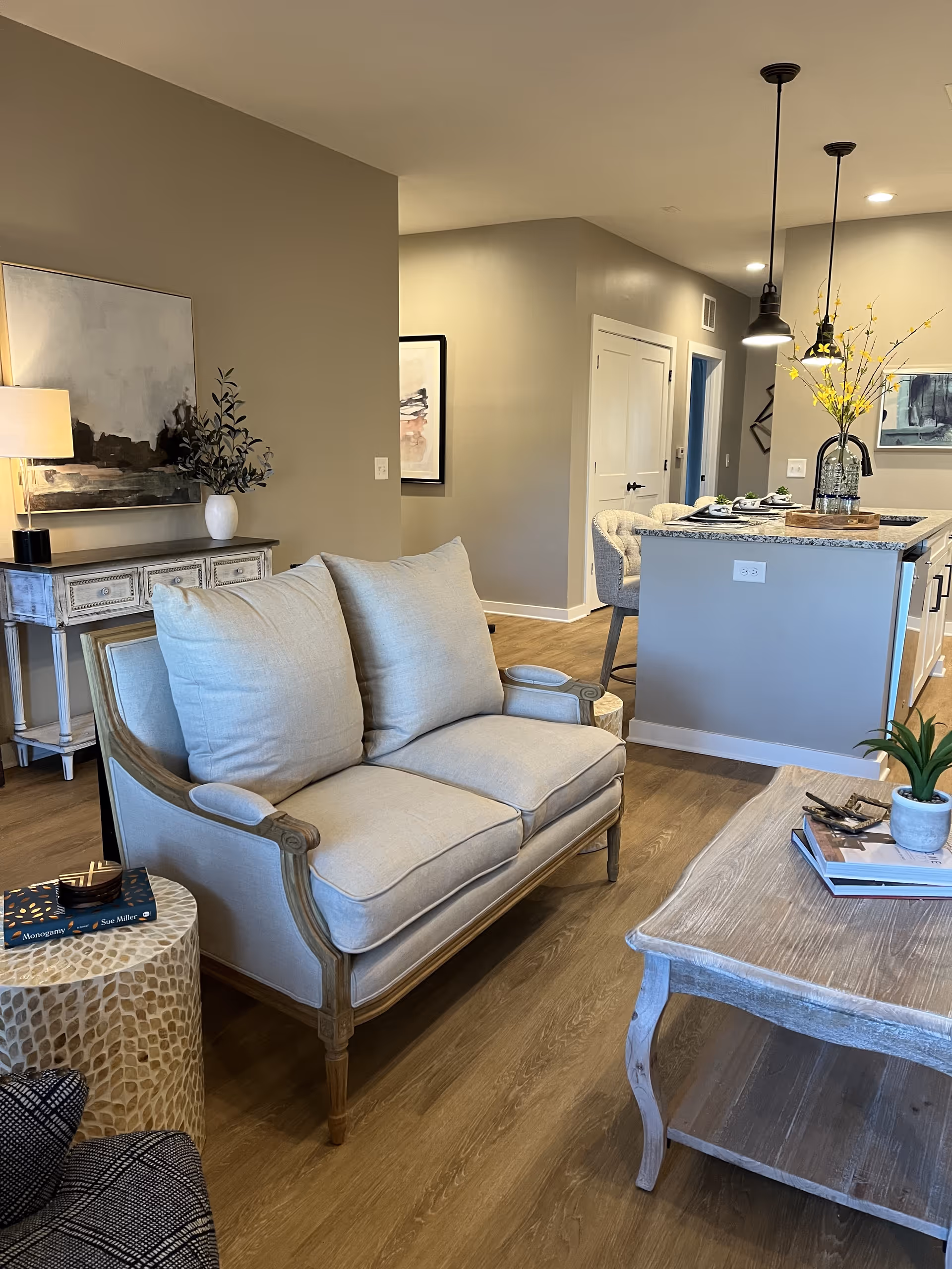 A cozy living room area with a beige cushioned loveseat, a wooden coffee table with books and a small plant, and a side table with a book on it. In the background, there is a kitchen island with bar stools, pendant lights, and decorative flowers in a vase. The walls are painted light beige, and there is a console table with a lamp and artwork on the wall.