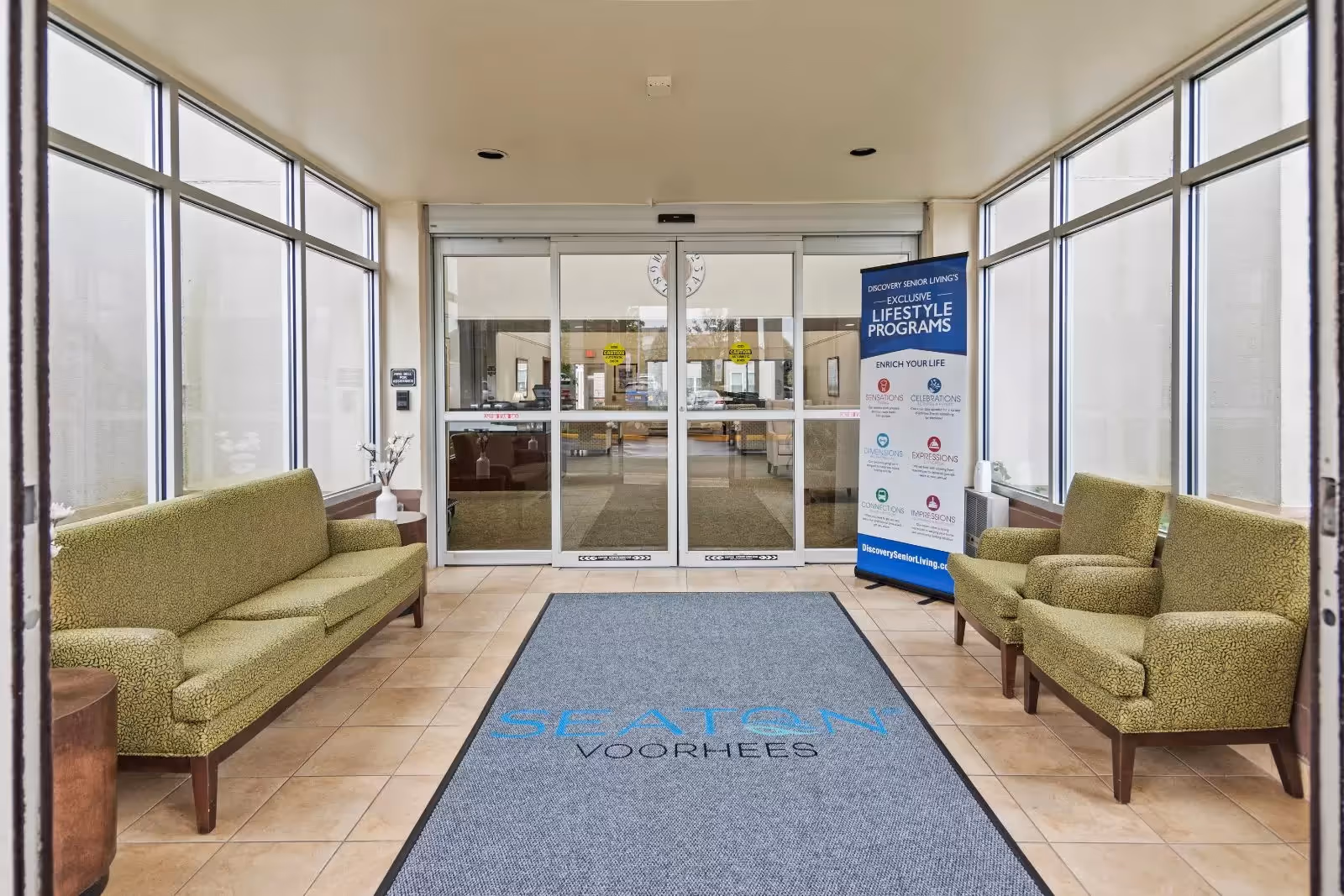 Entrance lobby of Seaton Voorhees facility with large glass windows on both sides, green upholstered seating on the left and right, a gray floor mat with the facility name printed on it, and automatic sliding glass doors at the far end. A standing banner on the right side displays information about lifestyle programs offered.