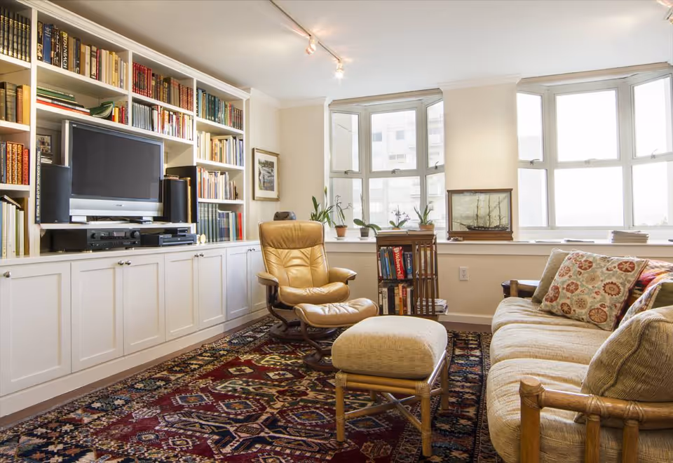 A cozy living room with a large built-in white bookshelf filled with books and a TV in the center. There is a tan leather recliner with a matching ottoman, a beige sofa with patterned cushions, a colorful patterned rug, and a bay window with several potted plants and a model ship on the windowsill.