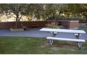 Outdoor seating area with a white picnic table on grass, brick planter walls, wooden benches and trees in the background.