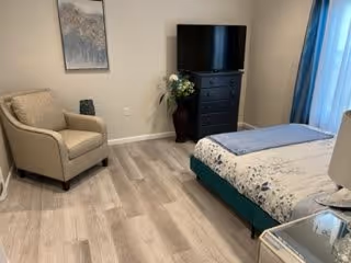 A bedroom with a bed covered in white and blue bedding, a beige armchair, a black dresser with a flat-screen TV on top, a vase with flowers next to the dresser, and a framed picture on the wall. The room has light wood flooring and a window with blue curtains.