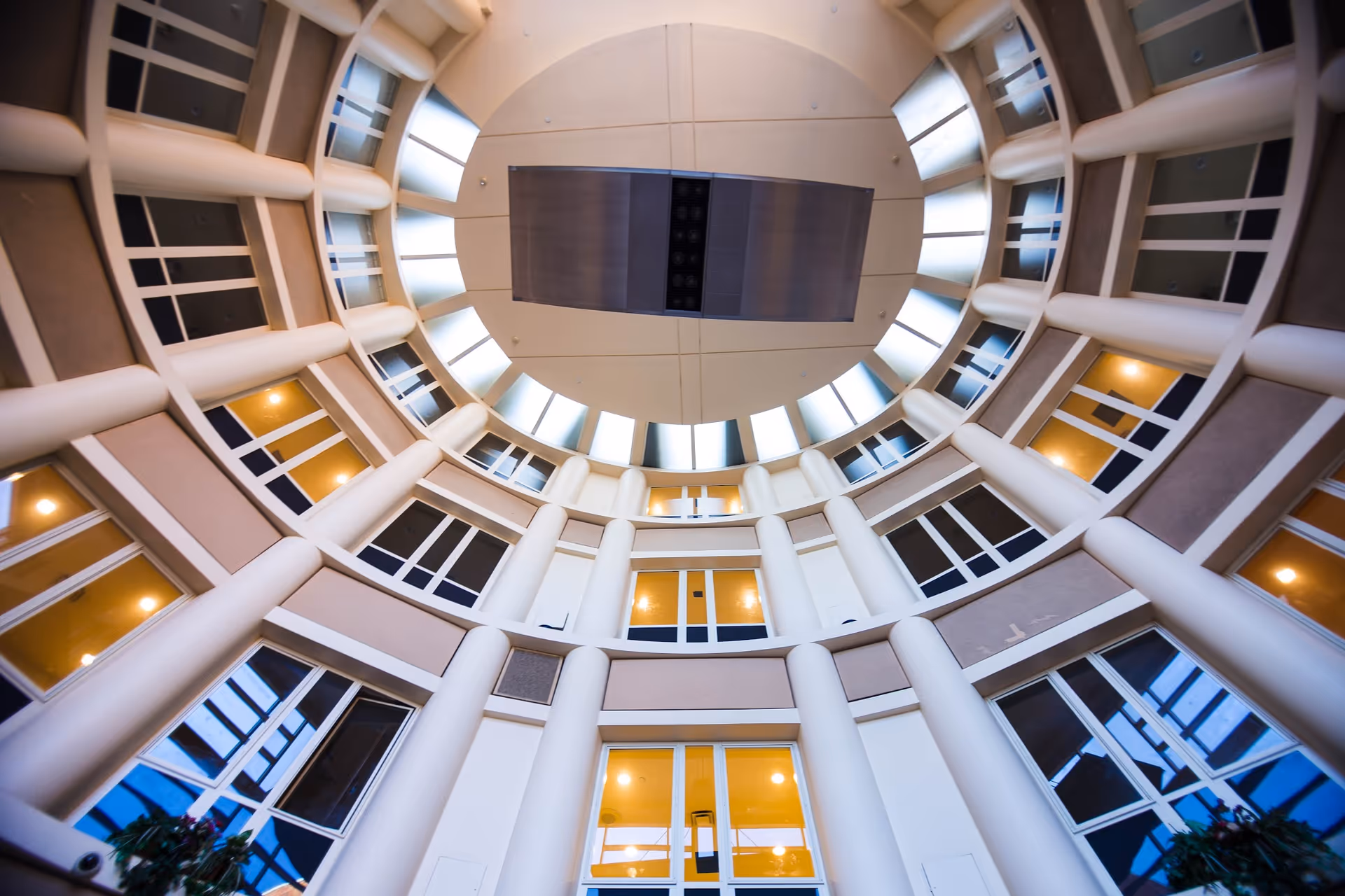 Interior view looking up at a circular atrium with multiple floors, large white columns, and windows surrounding the space. The ceiling features a circular skylight with a central rectangular fixture.