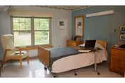 A simple, clean nursing facility bedroom with a single bed covered in white and blue bedding, a wooden chair near a window with a view of greenery outside, a wooden dresser with a mirror, and a laptop on a tray table over the bed.