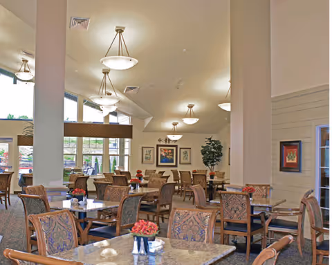 A spacious dining room with multiple tables and chairs arranged neatly. Each table has a small floral centerpiece. Large windows on the left side allow natural light to fill the room. The ceiling has several hanging light fixtures, and framed artwork decorates the walls. There is a tall plant in the corner, adding greenery to the space.