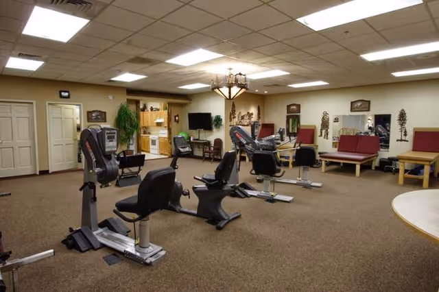 A spacious exercise room in a senior living facility with several stationary exercise bikes arranged on a carpeted floor. The room has beige walls with decorative wall hangings and mirrors. There are cushioned benches along the walls and a small kitchenette area with wooden cabinets and a TV in the background. The ceiling has recessed lighting and a chandelier-style light fixture.