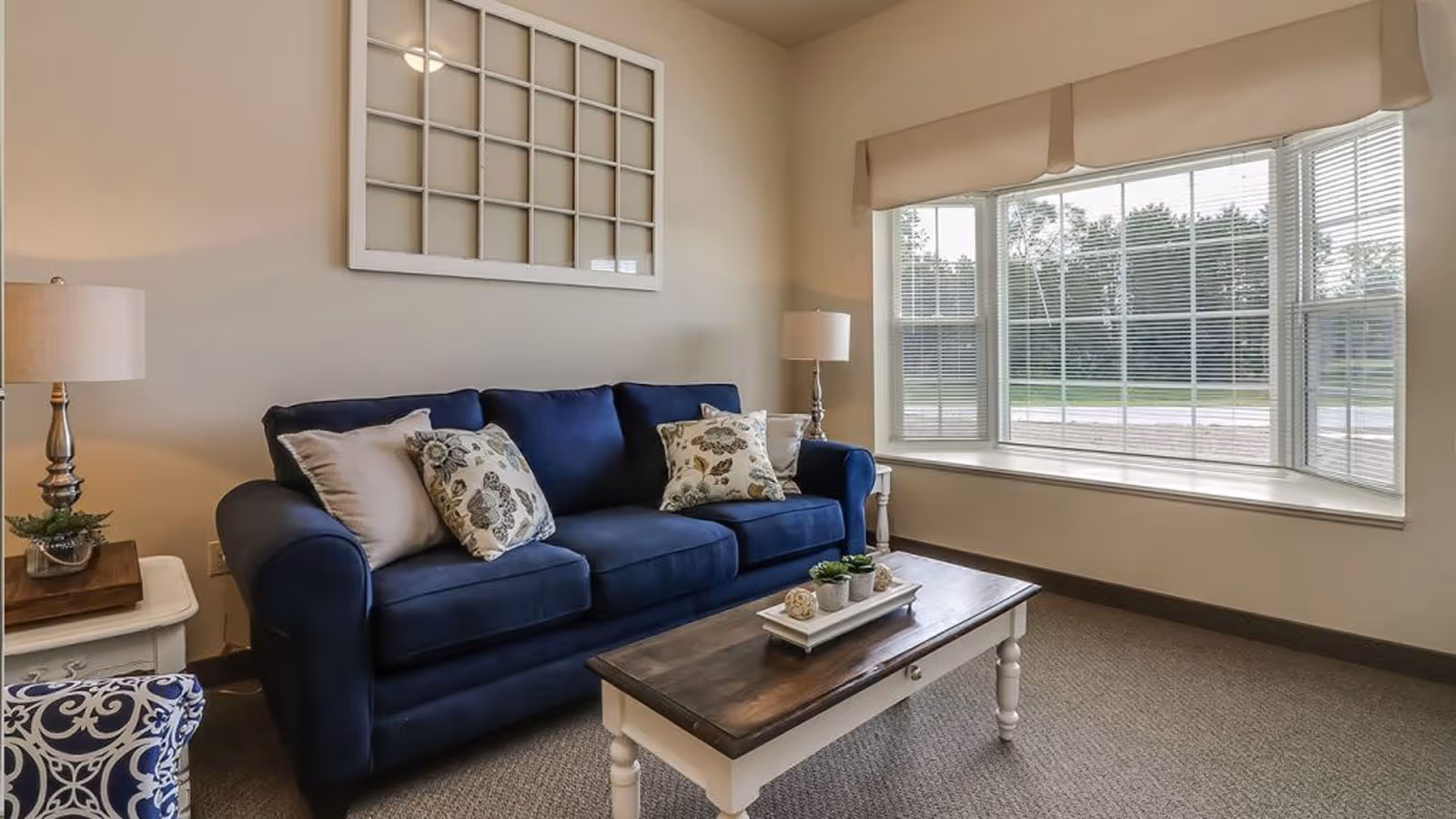 A cozy living room with a navy blue sofa adorned with floral and plain cushions, a wooden coffee table with small decorative plants, two side tables each with a lamp, and a large bay window with blinds letting in natural light. A decorative wall piece resembling a window frame hangs above the sofa.