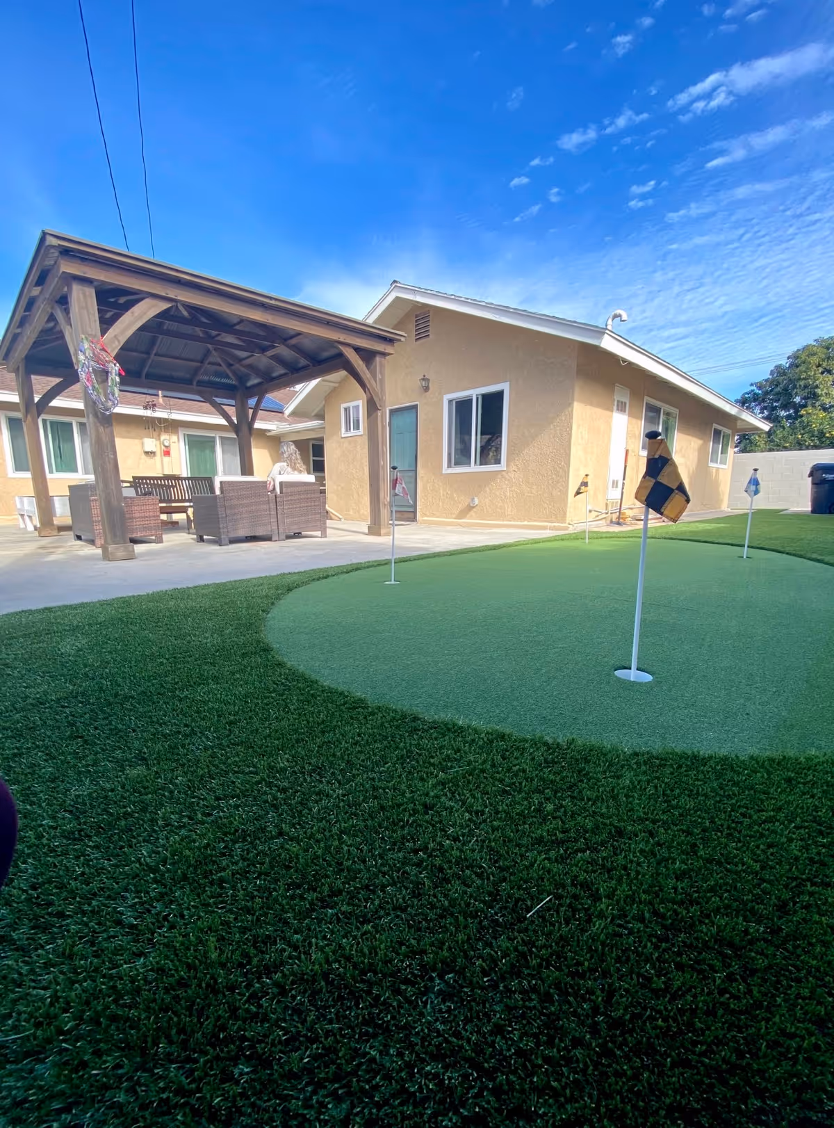 Outdoor area of a senior living facility with a small putting green featuring several golf holes with flags. There is a covered patio with wooden beams and outdoor seating including chairs and a bench. The building has beige walls and white-framed windows under a blue sky with some clouds.