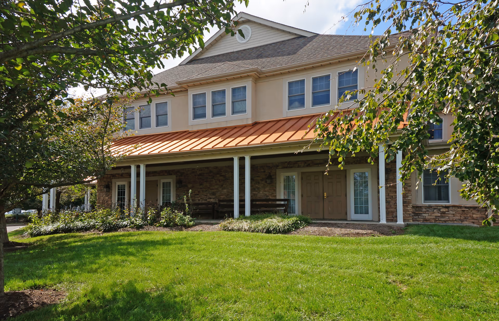 Exterior view of a two-story building with beige walls, multiple windows, a covered porch with white columns, and a green lawn in front. Trees partially frame the building on both sides.