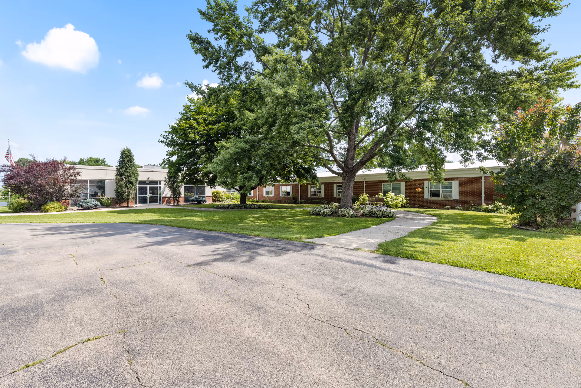 Exterior view of Franklin Grove Living and Rehabilitation Center showing a single-story brick building with large windows, surrounded by green grass, trees, and a paved driveway under a blue sky with some clouds.