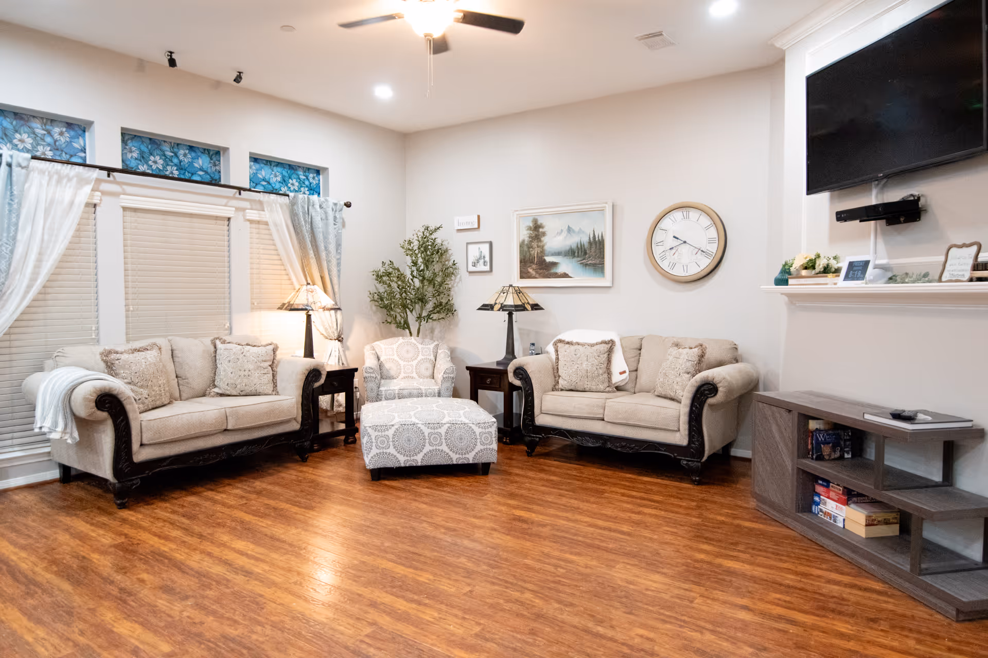 Communal living room with two beige sofas, a patterned ottoman, side tables and a wall-mounted TV over a mantel.