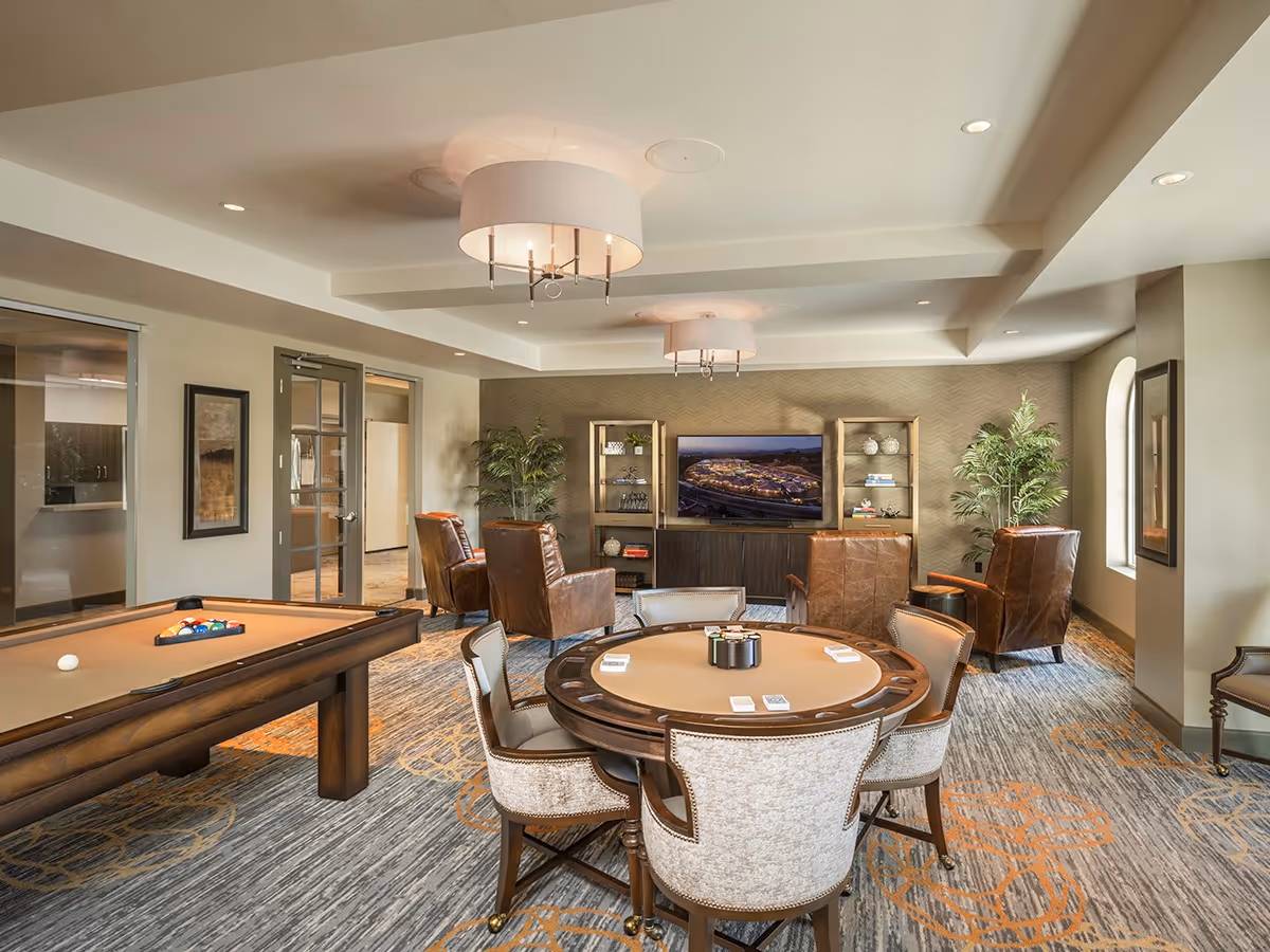 A well-lit common room with a pool table on the left and a round card table with six upholstered chairs in the center. In the background, there are four brown leather armchairs arranged around a TV mounted on a wall with shelves on either side. The room has large windows, plants, framed artwork, and modern ceiling light fixtures.