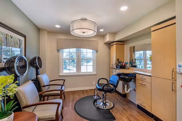 Interior view of a hair salon area in an assisted living facility with a styling chair on a black mat, two hair dryers on stands, two cushioned waiting chairs, a large mirror, wooden cabinets, and a window with a beige valance letting in natural light.