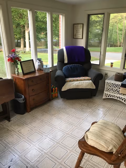 A cozy sitting area with a black armchair draped with a purple blanket and a cushion, positioned near large windows showing a green outdoor view. Next to the armchair is a wooden chest of drawers with a vase of flowers, framed text, and a small decorative item. A wooden chair with a cushion is partially visible in the foreground, and a white wicker side table with a small plant is on the right. The floor has a patterned tile design.