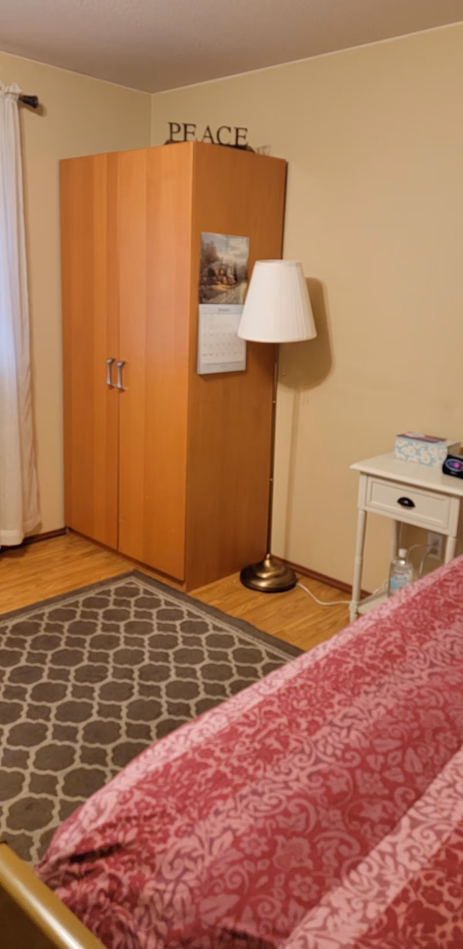 A bedroom corner with a wooden wardrobe, a standing lamp, a small white nightstand with a tissue box and clock, a patterned rug on the wooden floor, and part of a bed with a red patterned bedspread. The wall is beige with the word 'PEACE' partially visible above the wardrobe.