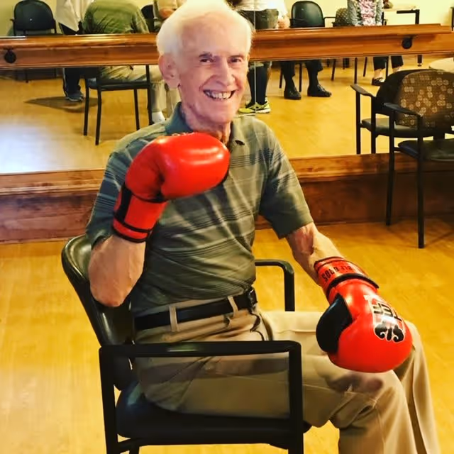 An elderly man sitting on a chair in a room with wooden floors and a large mirror on the wall behind him. He is smiling and wearing red boxing gloves, a green striped polo shirt, and beige pants. There are other chairs and people reflected in the mirror.