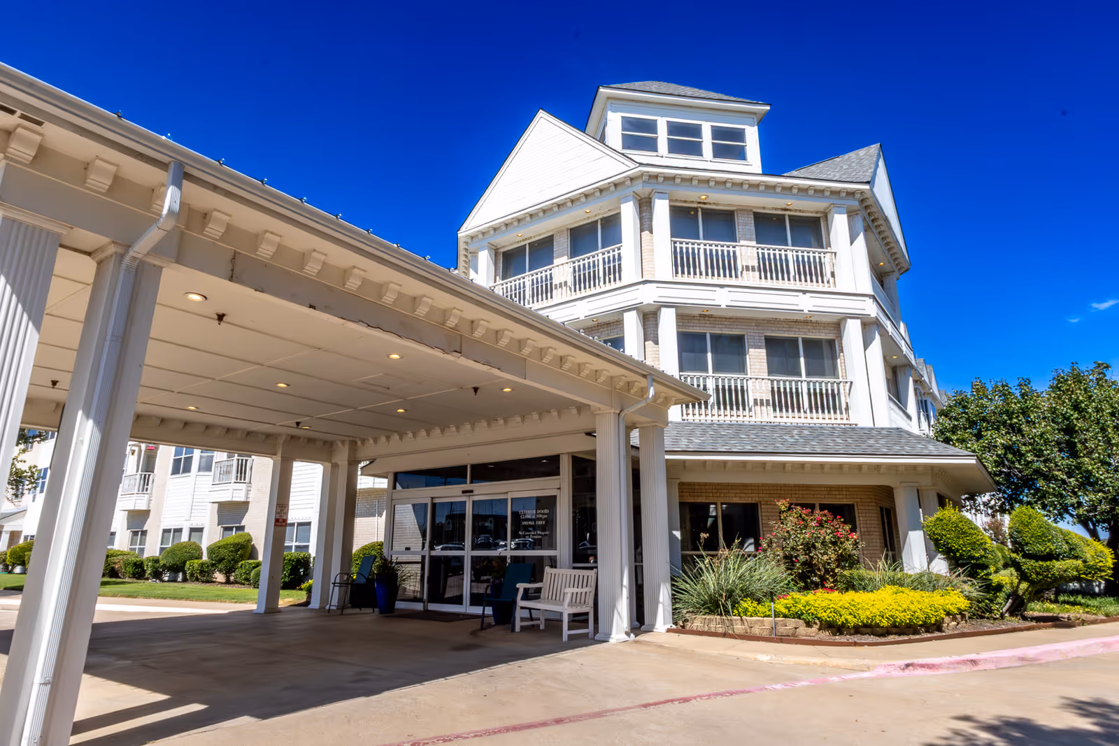 Exterior view of Lakestone Terrace Senior Living facility showing a multi-story building with white balconies and a covered entrance area. The sky is clear and blue, and there are well-maintained shrubs and greenery around the entrance.