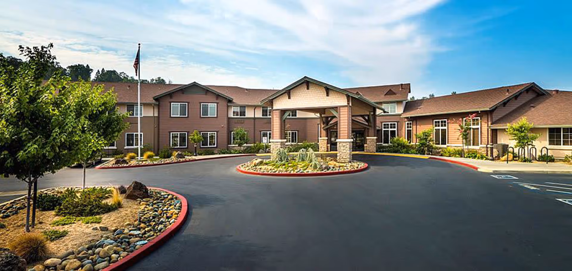 Front exterior view of Eskaton Village Placerville building with a covered entrance, landscaped roundabout with rocks and plants, trees, and an American flag on a flagpole under a partly cloudy sky.