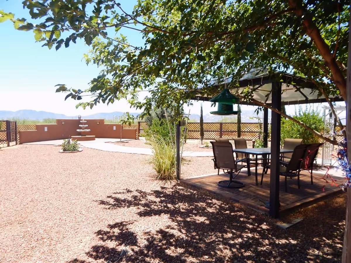 Outdoor garden area with a shaded gazebo containing a table and four chairs. There is a bird feeder hanging from the gazebo roof. The ground is covered with reddish gravel, and there are some plants and a multi-tiered water fountain in the background. Trees provide partial shade, and a wooden lattice fence encloses the area with mountains visible in the distance under a clear blue sky.