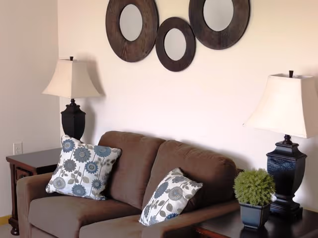A cozy living room area with a brown sofa adorned with two floral-patterned pillows. On either side of the sofa are dark wooden side tables, each holding a lamp with a white lampshade. A small green potted plant is on the right side table. Above the sofa, three round decorative mirrors with dark wooden frames are mounted on a light-colored wall.
