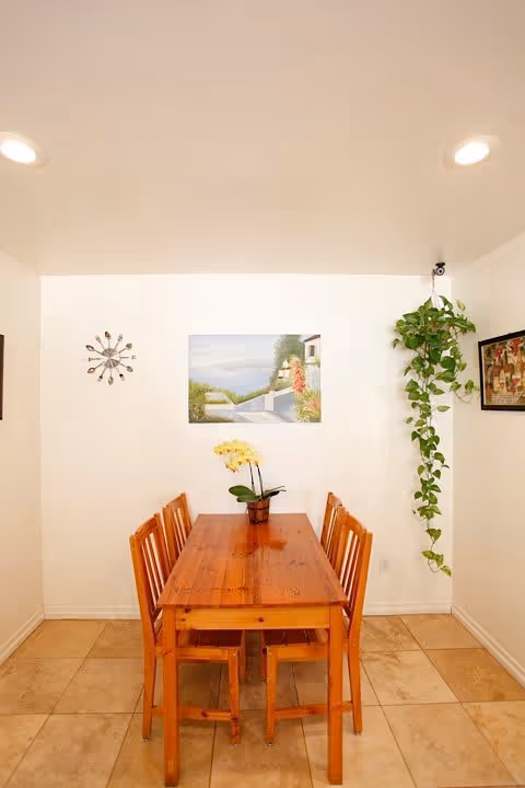 A small dining area with a wooden table and six matching chairs. A potted plant with yellow flowers is placed on the table. The walls are white and decorated with a clock, a framed painting of a scenic outdoor view, and a framed picture. A hanging green plant is suspended from the ceiling in the corner. The floor is tiled with beige tiles.