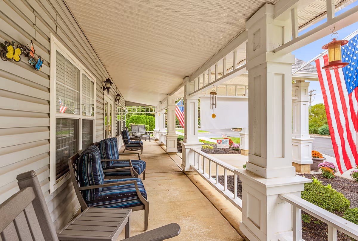 Covered porch area at Summit Place with several cushioned chairs and benches arranged along the wall. The porch has white pillars and railings, decorative butterfly wall hangings, and American flags displayed. There are wind chimes hanging from the ceiling and landscaped garden beds with flowers visible beyond the porch.