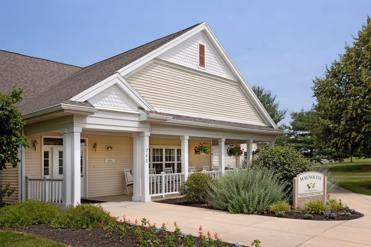 Exterior view of the Magnolias of Chambersburg senior living facility showing a beige building with white trim, a covered porch with rocking chairs, hanging flower baskets, and well-maintained landscaping including bushes and flowers under a clear blue sky.