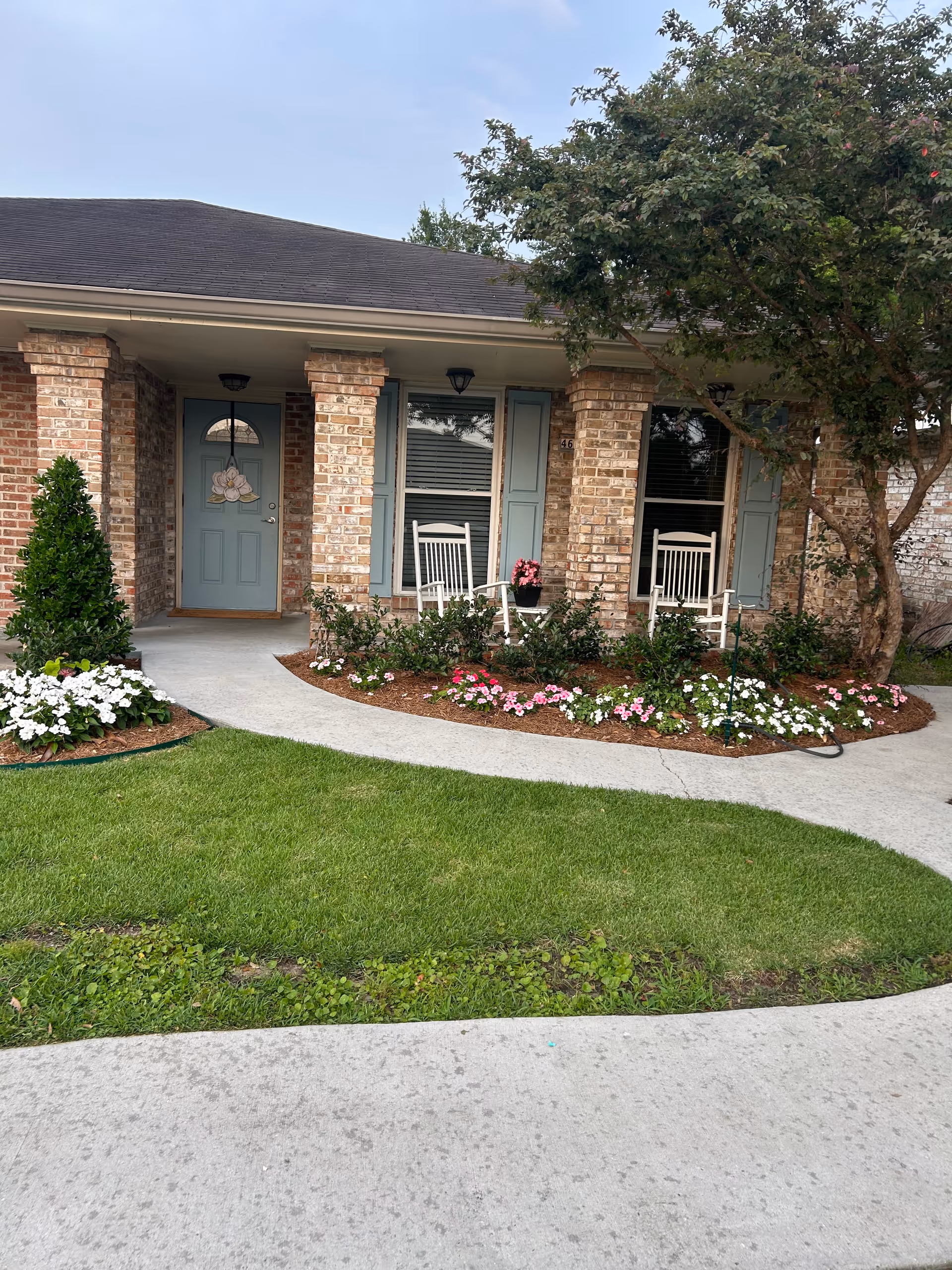 Front exterior view of a brick residence with a covered porch featuring two white rocking chairs, a blue door with a floral decoration, flower beds with white and pink flowers, a tree, and a well-maintained lawn with a curved concrete walkway.