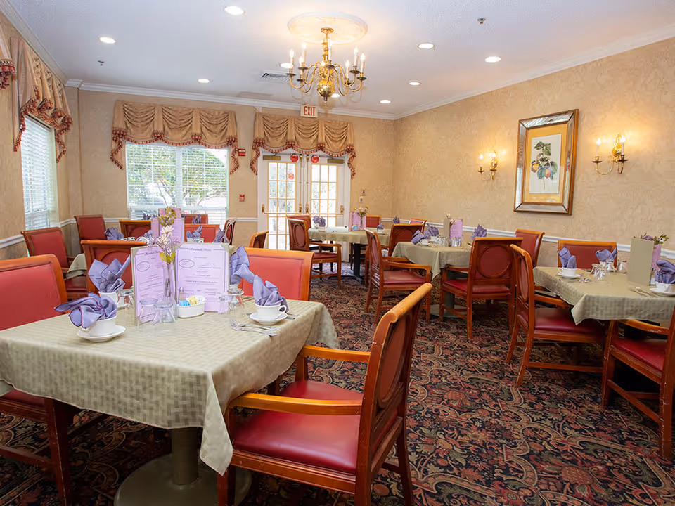Dining room with tables set with folded purple napkins, menus, china, and red upholstered chairs under chandeliers.